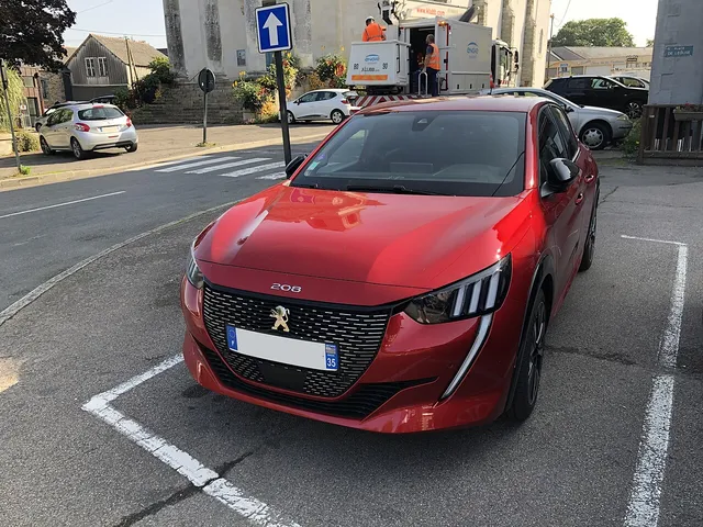Peugeot 208 rouge de face, garée sur une place de village